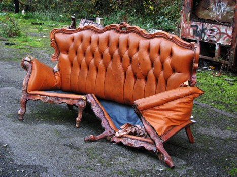 A vintage-style sofa with a wooden carved frame and upholstered in brown leather or faux leather fabric, featuring a tufted backrest and rolled arms. The sofa is visibly damaged, with one side torn open showing the internal cushioning and springs, and the fabric on the armrest appears crumpled and worn. It is placed outdoors on a paved surface, with a garden or park area in the background, including trees and greenery. The setting suggests it is part of rubbish or debris collection, possibly being prepared for removal by a waste management company such as Commercial Waste Removal Kensington. The lighting indicates a daytime scene, with natural light illuminating the scene, highlighting the textures of the upholstery, the intricacies of the wooden frame, and the surrounding environment. The photograph captures the sofa in the process of being cleared or disposed of, fitting within the context of rubbish removal services in Kensington or nearby areas.