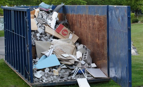 A large industrial metal skip positioned outdoors on a grassy area, filled with a mixture of construction waste including broken drywall, plasterboard, cut wood, cardboard boxes, and discarded tiles, with some debris spilling over the top edge onto the ground. The skip has a dark blue exterior with a partially rusted interior surface, situated on a patch of green grass with trees visible in the background, indicating a typical urban or suburban environment. The image captures the scene during daytime with natural lighting highlighting the textures of the various waste materials, supporting professional rubbish removal services such as those provided by Commercial Waste Removal Kensington in the locality near postcode W14, serving the Kensington area in London.