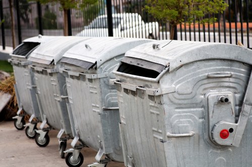 Four large outdoor metal wheelie bins with hinged lids, lined up side by side on a paved surface in an urban setting, with a black metal fence and some greenery in the background. The bins are made of galvanized steel with a slightly textured, silver-grey finish, each featuring two wheels at the front for mobility and a small locking mechanism on the side. The lids are closed, with slight variations in angle, and the bins appear to be used for general rubbish collection. The environment suggests a commercial or residential waste disposal area, possibly in Kensington or nearby London, as indicated by the website context. The lighting is natural, illuminating the scene evenly, and the overall image reflects typical rubbish removal infrastructure managed by companies like Commercial Waste Removal Kensington.