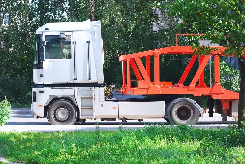 A white commercial truck with a black bumper and side paneling is parked on a paved road in a green residential area. The truck features a passenger cab with a large front windshield, side mirror, and visible door handles. Attached to the back of the truck is an open, orange metal framework designed to hold waste or bulky items, with vertical and diagonal supports forming a sturdy structure. The framework is empty and sits on a flatbed base, positioned adjacent to a patch of well-maintained grass and a background of leafy trees and shrubs, with some buildings partially visible behind the foliage. The scene is illuminated by natural daylight, indicating a clear day, and the overall setting aligns with a rubbish removal service operating within a suburban environment, possibly near Kensington or a similar London district. This image is relevant to services offered by Commercial Waste Removal Kensington, focusing on the transportation and disposal of rubbish materials.