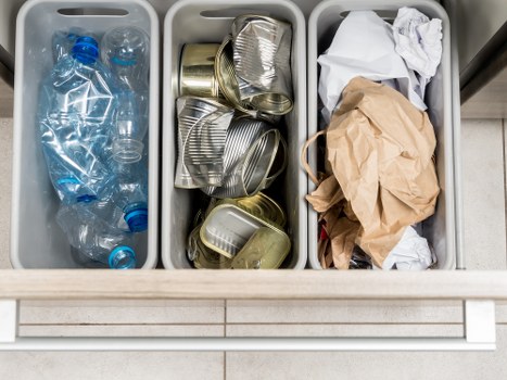 A close-up view of three grey plastic recycling bins placed side by side on a tiled floor, each filled with different types of waste. The left bin contains several crumpled plastic water bottles with blue caps, partially crushed, and transparent plastic packaging. The middle bin holds a variety of aluminium cans, some flattened and others slightly rounded, with visible ridges and metallic sheen. The right bin contains crumpled paper bags and used tissue paper, mostly brown kraft and white, with a mix of textures from smooth to crinkled. The bins are positioned outdoors on a paving surface, with a neutral background suggesting a residential or commercial waste collection area. The arrangement clearly reflects waste segregation efforts typical of rubbish removal services, such as those provided by Commercial Waste Removal Kensington, serving locations around Kensington and the W14 postcode area.